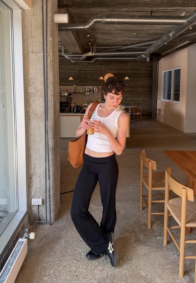 Mujer joven con top blanco sin mangas y pantalones negros sosteniendo un café con hielo, de pie en una cafetería moderna con muebles de madera y tuberías expuestas.