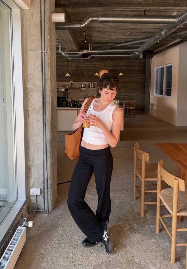 Mlada žena u bijeloj majici bez rukava i crnim hlačama drži ledenu kavu, stojeći u modernom kafiću s drvenim namještajem i izloženim cijevima.