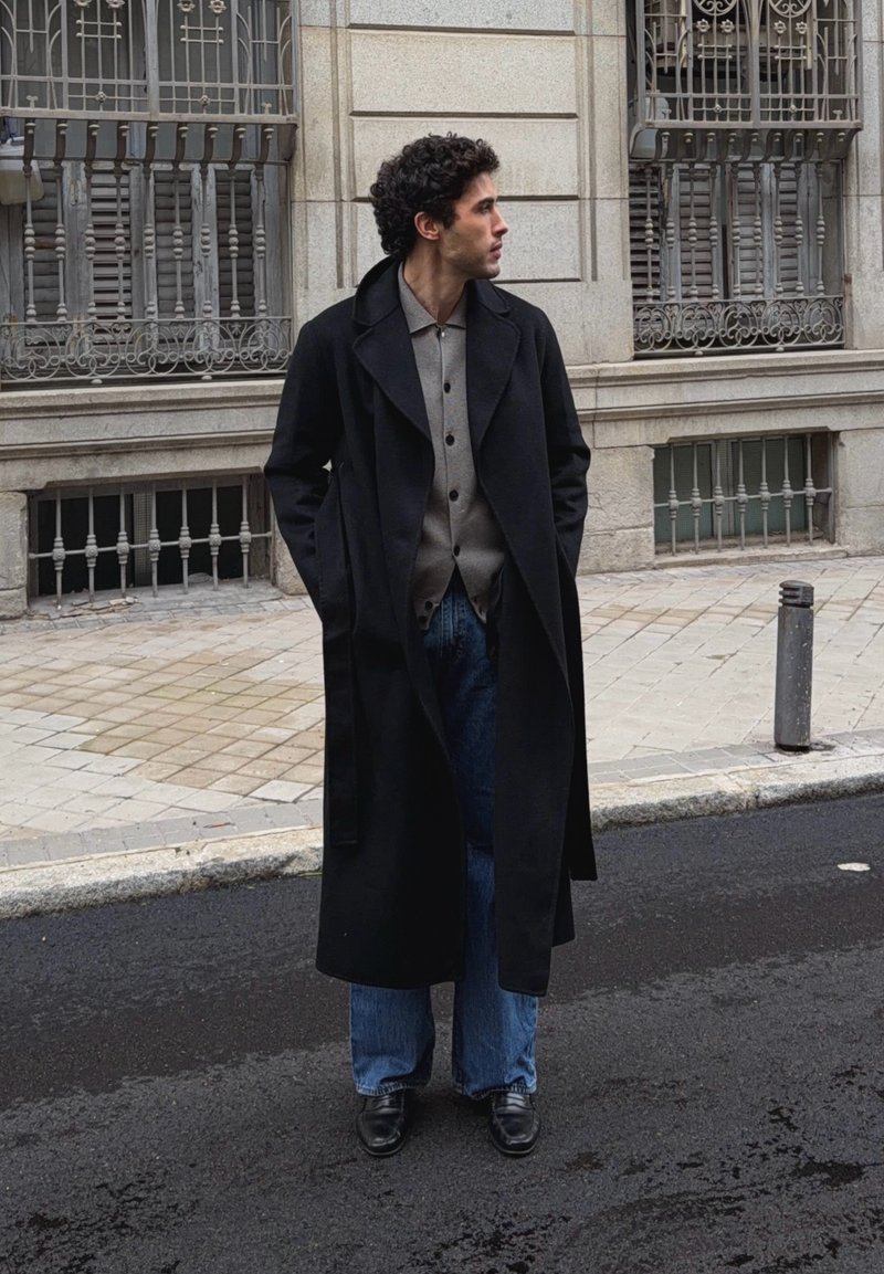 Homme aux cheveux bouclés portant un long manteau noir, une chemise grise, un jean bleu et des chaussures noires, debout dans la rue à côté d'un bâtiment en pierre avec des fenêtres en métal.