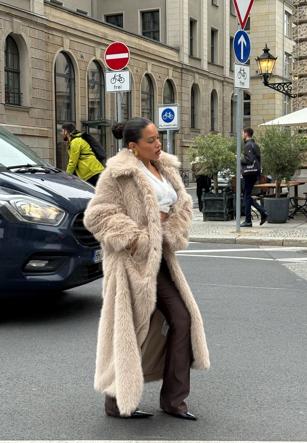 A woman in a long beige fur coat and brown trousers stands on a city street next to bicycle traffic signs and a blue car.