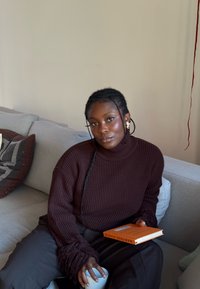 Pull à col roulé côtelé marron avec détails en franges, associé à un pantalon large sombre. Tenant une tasse blanche et un livre orange sur un canapé gris.