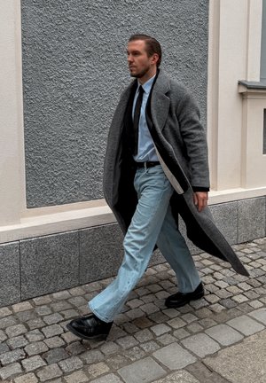 Hombre caminando por una calle empedrada con un abrigo gris, camisa azul claro, corbata negra, vaqueros azul claro y zapatos negros.