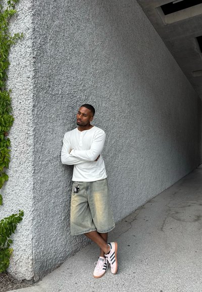 Hombre con camisa de manga larga blanca y pantalones cortos desgastados hasta la rodilla se apoya contra una pared texturizada gris, con llaves sujetas a sus pantalones cortos.