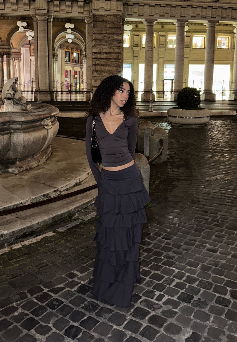 Femme portant une longue jupe noire à étages et un haut noir, debout dans une rue pavée près d'une fontaine en pierre éclairée et des arches la nuit.