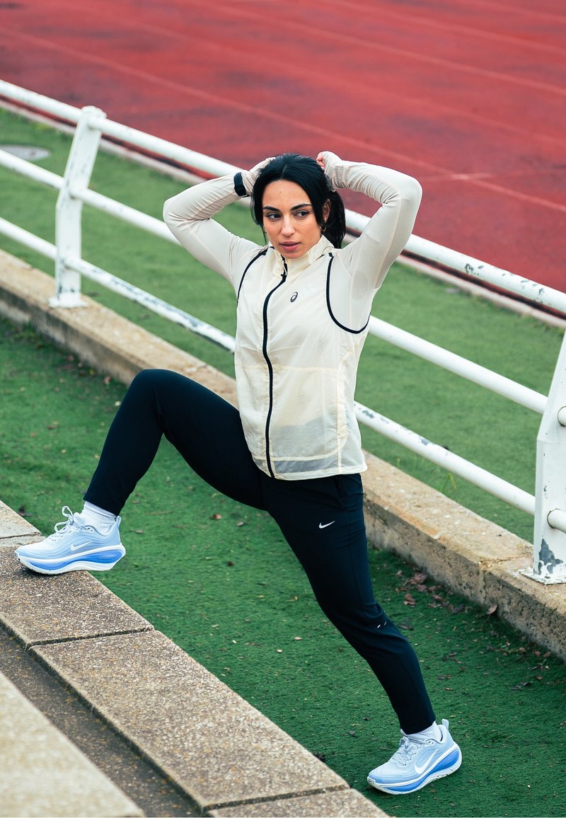 Veste légère crème à fermeture éclair avec bordure noire, pantalon de sport noir et chaussures de course Nike bleues. Entraîneur en position d'étirement sur une piste.