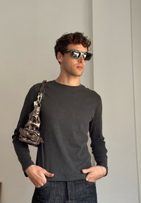 Grey long-sleeve shirt, dark denim jeans, and rectangular sunglasses. A textured metallic shoulder bag hangs from the left shoulder.