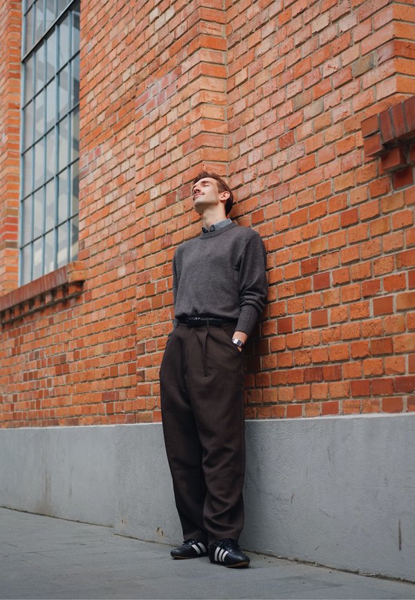 Grijze trui, bruine high-waisted broek, zwarte schoenen met witte strepen, staand tegen een gebakken stenen muur in textuur. Horloge om de pols.