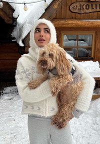 Fehér, bolyhos cipzáras kapucnis dzsekit viselő nő egy világosbarna kutyát tart, amely szürke pulóvert visel, havas, fából készült háttér előtt.