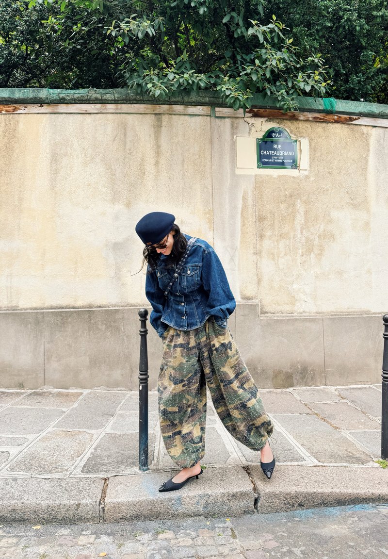 Femme portant une veste en jean, un pantalon camouflage, un béret et des lunettes de soleil, s'appuyant sur un poteau noir sur un trottoir en pierre près d'un mur beige.