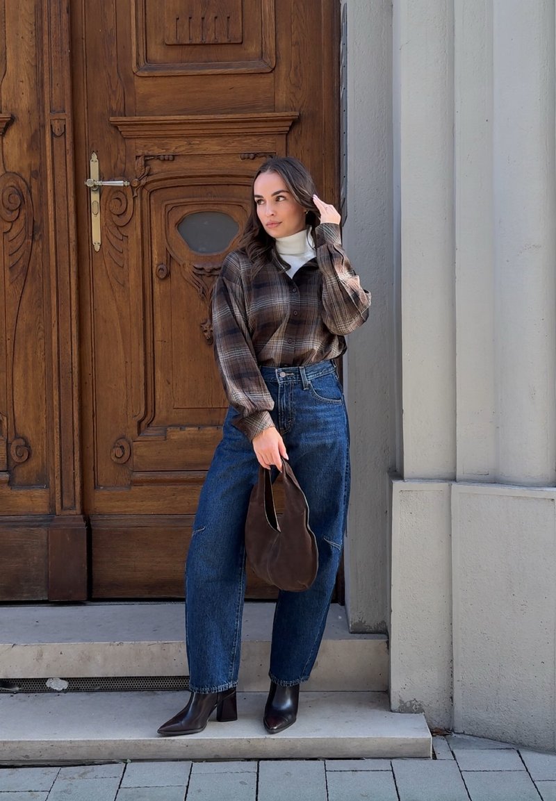 Femme debout sur des marches en pierre près d'une grande porte en bois ornée, portant une chemise à carreaux, un jean taille haute, des bottes noires, tenant un sac marron et se touchant les cheveux.