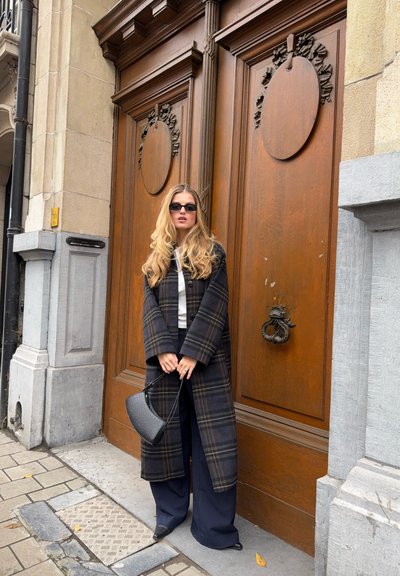 Abrigo largo de cuadros en colores oscuros sobre una camiseta blanca, combinado con pantalones anchos azul marino y sosteniendo un bolso negro. Puerta de madera y pared texturizada.