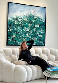 Γυναίκα με μαύρο φόρεμα που ξαπλώνει σε λευκό καναπέ με κουμπιά κάτω από μεγάλο αφηρημένο ζωγραφικό έργο σε μπλε-πράσινους τόνους, δίπλα σε μαρμάρινο τραπεζάκι καφέ με στοιβαγμένα βιβλία.
