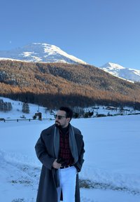 Manteau gris par-dessus une chemise à carreaux rouge, pantalon blanc et écharpe grise. Sol et montagnes couverts de neige en arrière-plan sous un ciel bleu clair.