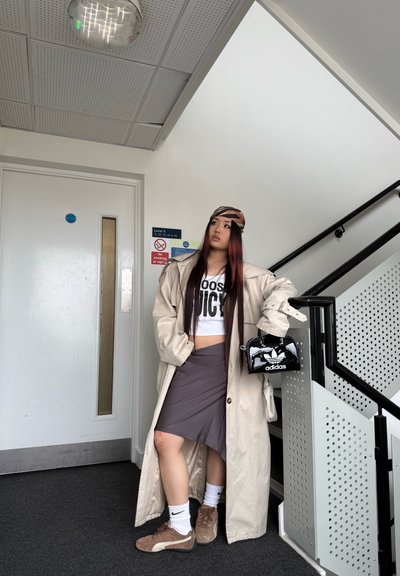 Abrigo largo beige sobre un top corto blanco con texto "Juicy", falda gris y zapatillas marrones. Bolsa negra de Adidas, de pie cerca de una escalera.