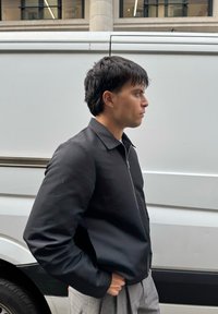 Crna jakna na patent s ovratnikom, nošena preko svijetle košulje, u kombinaciji sa sivim prugastim hlačama. Opusten ambijent na ulici.