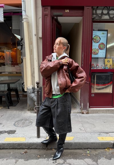 Joven mujer con chaqueta de cuero marrón, sudadera con capucha gris, pantalones cortos anchos negros y botas puntiagudas negras de pie en una acera de la ciudad.