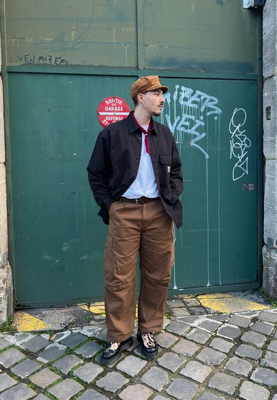 Pantalones cargo marrones, camisa negra, camiseta blanca con cuello rojo, gorra marrón, zapatos de plataforma negros con acentos estampados, de pie contra una pared verde.