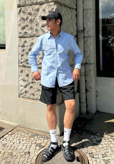 Camisa azul claro de botones, pantalones cortos negros, calcetines blancos y sandalias negras. Fondo de pared texturizada, día soleado. Atuendo casual con un corte cómodo.