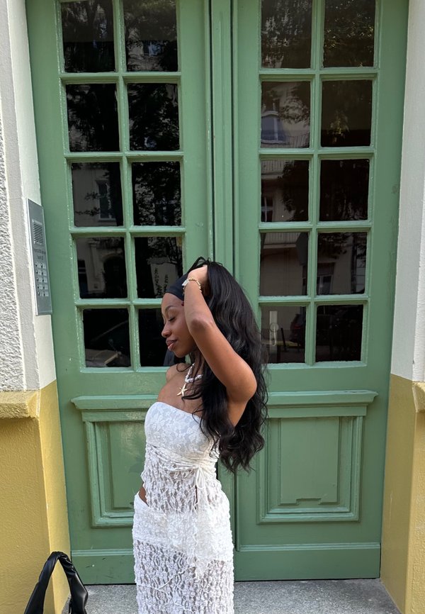 White lace dress with spaghetti straps, features a side tie detail, textured pattern, worn against a green door backdrop. Black bag at the feet.