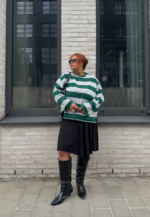 Persona con cabello rojo corto, que lleva gafas de sol negras, suéter a rayas verdes y blancas, falda negra y botas negras hasta la rodilla, de pie junto a una pared de ladrillos.