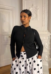 Cardigã preto de mangas longas canelado com colarinho e detalhes de botões, combinado com uma saia branca com grandes bolinhas pretas.