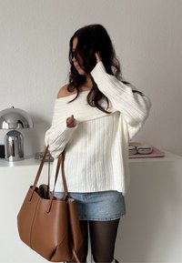 Femme ajustant ses cheveux, portant un pull blanc à épaules dénudées, une jupe en denim, des collants noirs, tenant un grand sac à main en cuir marron à l'intérieur près d'une armoire blanche.