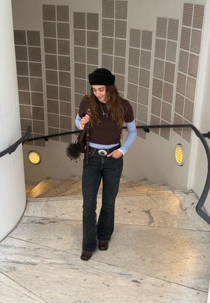 Mujer joven con boina negra, mangas largas en capas, jeans oscuros y botas marrones, de pie en una escalera de mármol con barandilla de metal en el interior.