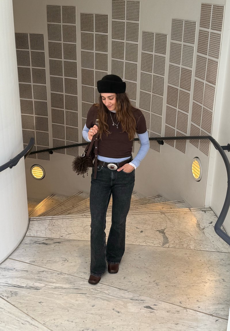 Νεαρή γυναίκα που φοράει μαύρο μπερέ, στρωματοποιημένα μακριά μανίκια, σκουρόχρωμο τζιν και καφέ μπότες, στέκεται σε μαρμάρινη σκάλα με μεταλλική κουπαστή σε εσωτερικό χώρο.