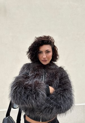 Mujer con cabello rizado y oscuro, vistiendo una chaqueta de piel negra y esponjosa, de pie contra una pared lisa de color gris claro con los brazos cruzados.