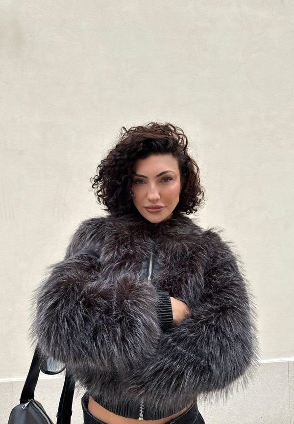 Femme aux cheveux bouclés et foncés portant une veste en fourrure noire et duveteuse, debout contre un mur gris clair uni, les bras croisés.