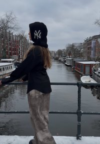 Personne portant un sweat à capuche noir avec un logo blanc "NY" et un pantalon gris, appuyée sur une rambarde surplombant un canal enneigé avec des bateaux et des bâtiments traditionnels.
