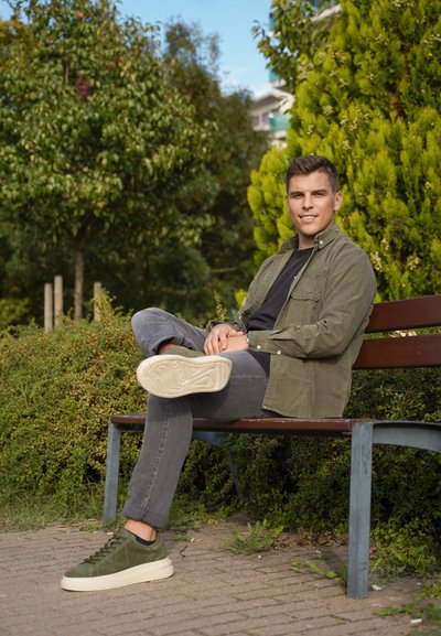 Camisa de botones verde oliva sobre una camiseta negra, jeans grises y zapatillas de ante verdes con suelas blancas. Sentado en un banco del parque.