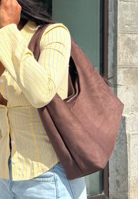 Bolso tote marrón con una textura suave y una abertura ancha, llevado al hombro, combinado con una camisa de botones a rayas amarillas.