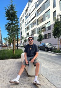 Jaunas vyras sėdi ant kreivų baltų betono suolelių, vilkėdamas juodą marškinėlį ir džinsinius šortus, su akinių nuo saulės ir juodai baltais sportbačiais.