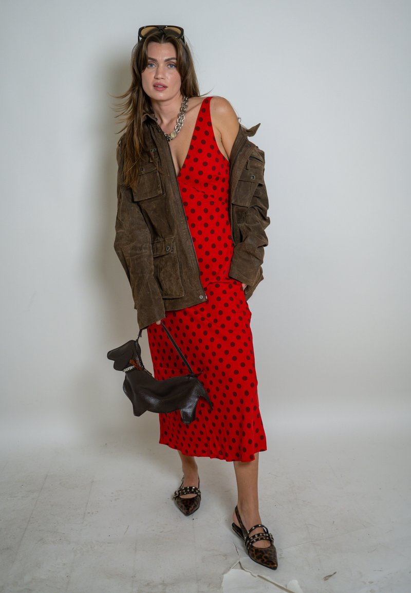 Donna con abito rosso a pois e giacca marrone scivolata sulle spalle, che tiene una borsa nera a forma di cane e indossa scarpe con stampa leopardata.