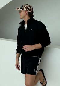 Άνδρας με καπέλο καμουφλάζ, μαύρο φούτερ Nike με φερμουάρ μέχρι τη μέση, σορτς Adidas και μαύρα σανδάλια, ακουμπώντας σε λευκό τοίχο και κοιτάζοντας πλαγίως.