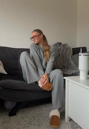 Suéter gris de punto y pantalones de pierna ancha, zapatillas marrones con detalles en crema, sentado en un sofá oscuro, con un vaso blanco cerca.