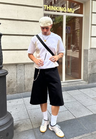 Camiseta blanca con un logo, pantalones cortos negros y holgados, calcetines blancos y zapatos grises con suelas naranjas. Los accesorios incluyen una cadena y collares en capas.