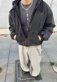 Personne portant une veste noire zippée, un pull côtelé gris, des colliers en argent superposés avec des croix, un pantalon de jogging gris clair avec une chaîne, et des chaussures noires.