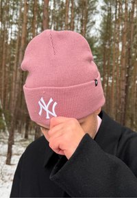 Personne en manteau noir ajustant un bonnet en tricot rose avec un logo NY blanc, debout dans une forêt enneigée avec de grands pins.