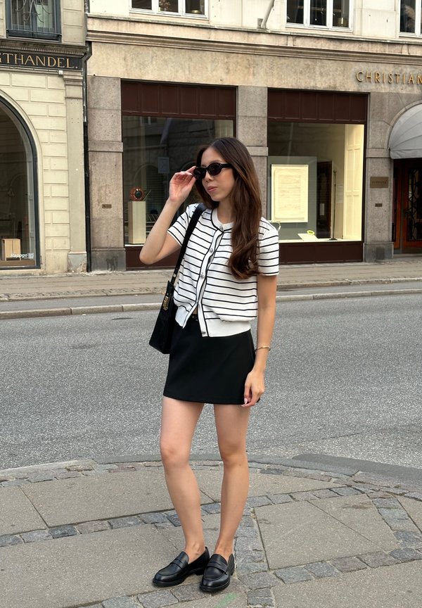 Mocasines negros, una falda negra y una blusa de punto blanca a rayas con mangas cortas. Unas gafas de sol negras y un bolso negro completan el conjunto.
