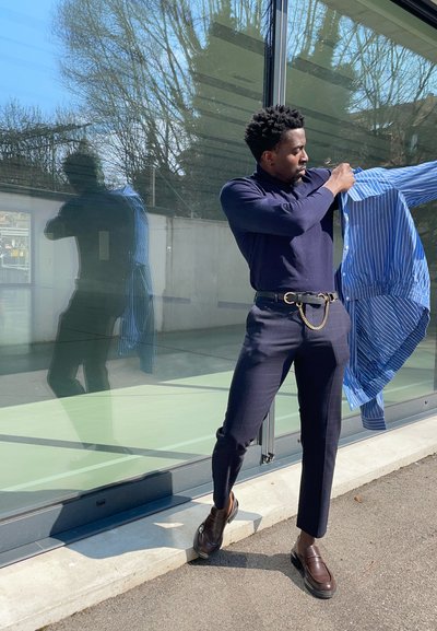 Camisa de manga larga azul marino, camisa rayada azul, pantalones oscuros a medida, cinturón negro con detalle de cadena y mocasines de cuero marrón. Fondo de cristal reflectante.