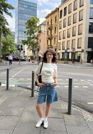 Mujer joven con cabello castaño vestida con una camisa blanca y shorts de mezclilla se encuentra en la acera de la ciudad cerca de un paso de cebra, con edificios y árboles al fondo.