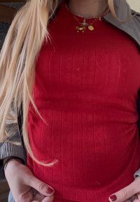 Longs cheveux blonds sur un pull rouge texturé avec des pendentifs en forme de crabe doré et de planète sur des chaînes. Les mains tiennent l'ourlet du pull, les ongles sont peints en rouge.