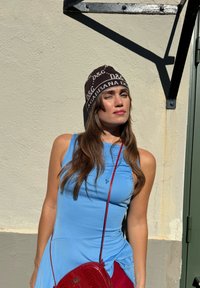 Gorro de malha castanho com impressão de logótipo branco, top sem mangas azul e mala de ombro vermelha feita de material texturizado, em fundo de parede clara.