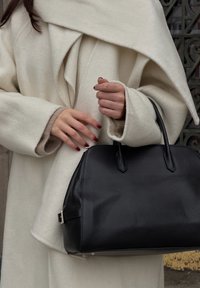 Črna usnjena torbica z dvema ročajema, predstavljena na svetli volneni jakni z ogrnjencem.