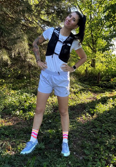 Chaleco de running ligero negro, camiseta blanca, pantalones cortos gris claro, zapatillas azules, calcetines de zigzag multicolores. Entorno al aire libre con vegetación.