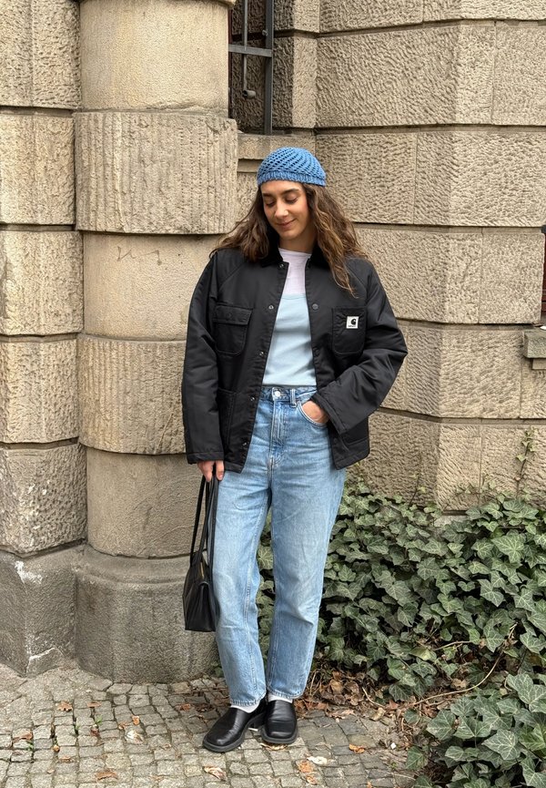 Ung kvinde i lyseblå hue, sort jakke, lyseblå jeans og sorte sko står ved en stenmursvæg dækket med grøn vedbend og holder en sort taske.