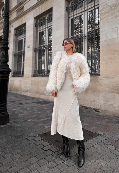 Abrigo de pelo sintético blanco y esponjoso sobre un vestido de canalé claro con un dobladillo asimétrico. Botines negros de punta completan el conjunto.