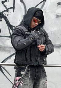 Μαύρο τζάκετ από τζιν με φθαρμένες άκρες και μεγάλα γρύλια. Φορεμένο πάνω από μια μαύρη φούτερ. Διαθέτει ασημένιες αλυσίδες και ροζ γκράφιτι σε τζιν παντελόνι.